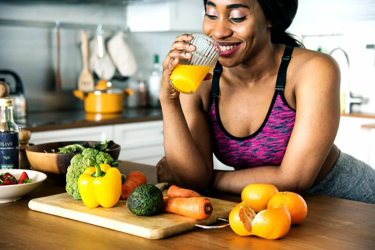 A woman drinking juice from a glass in front of vegetables.